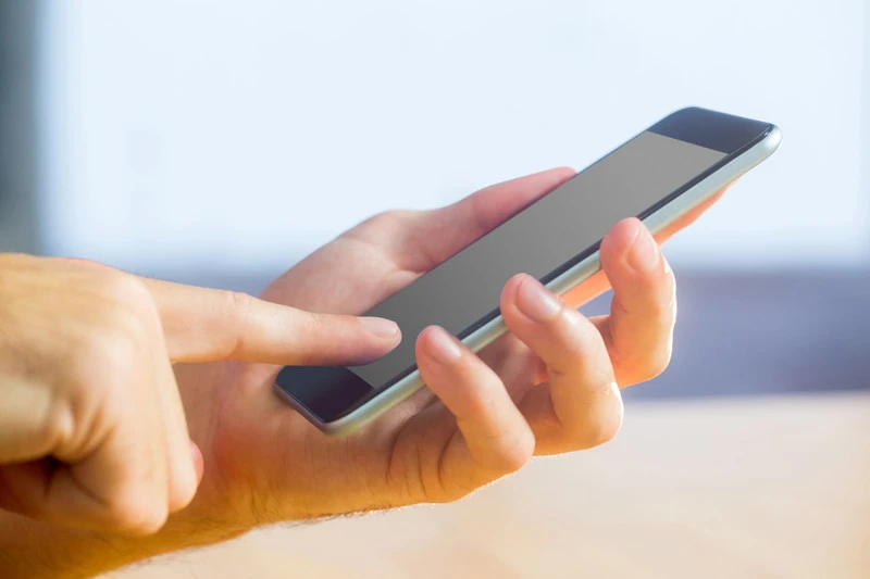 En närbild av en hand som håller i en smartphone medan pekfingret från den andra handen rör vid skärmen. Bakgrunden är suddig vilket gör att fokus ligger på telefonen och händerna.