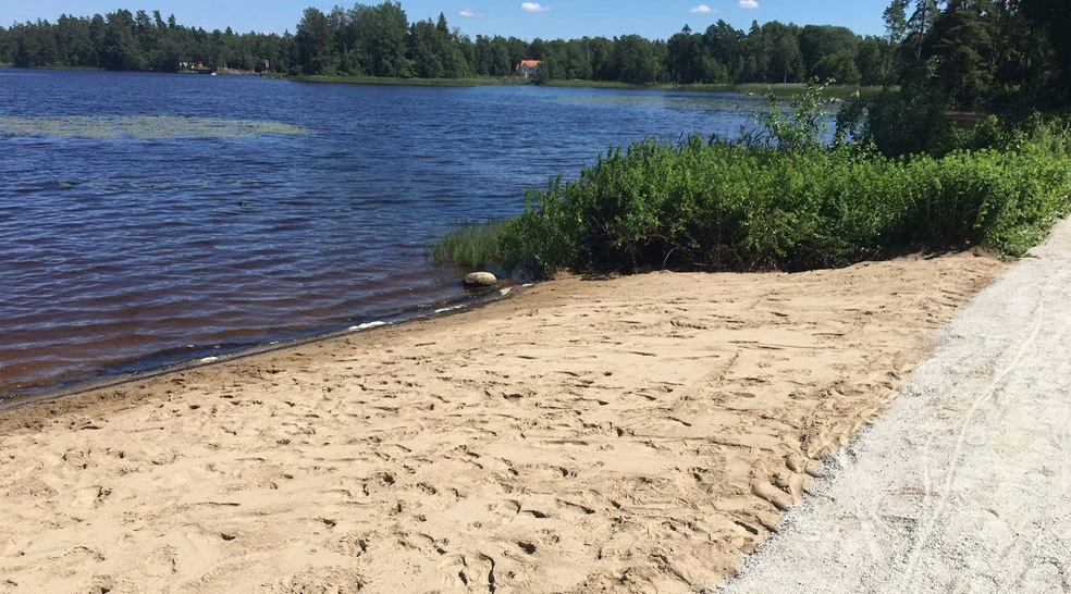 En sandstrand vid en sjö med blått vatten, omgiven av grönska. En grusväg löper längs stranden, och i bakgrunden syns skog samt ett hus med rött tak. Himlen är klar och blå, vilket skapar en somrig atmosfär.
