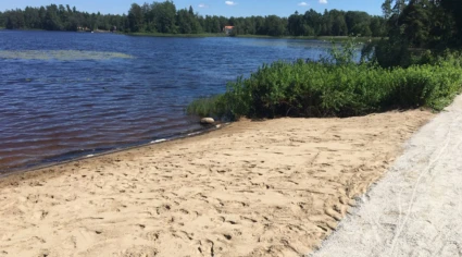 En sandstrand vid en sjö med blått vatten, omgiven av grönska. En grusväg löper längs stranden, och i bakgrunden syns skog samt ett hus med rött tak. Himlen är klar och blå, vilket skapar en somrig atmosfär.