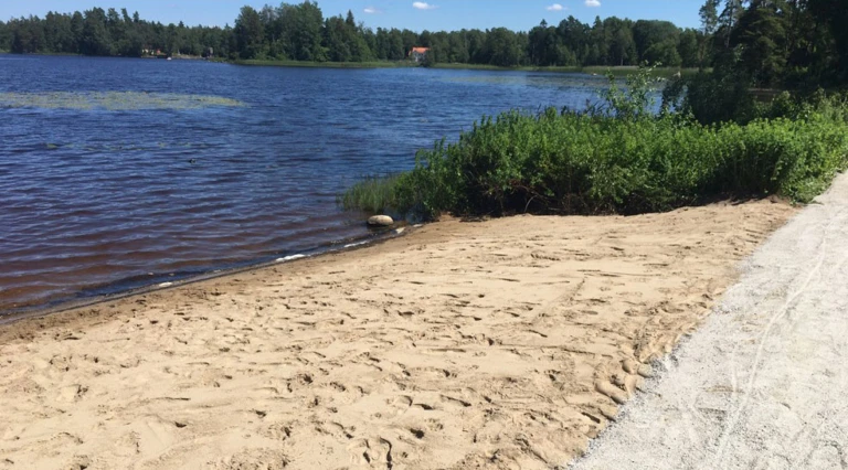 En sandstrand vid en sjö med blått vatten, omgiven av grönska. En grusväg löper längs stranden, och i bakgrunden syns skog samt ett hus med rött tak. Himlen är klar och blå, vilket skapar en somrig atmosfär.