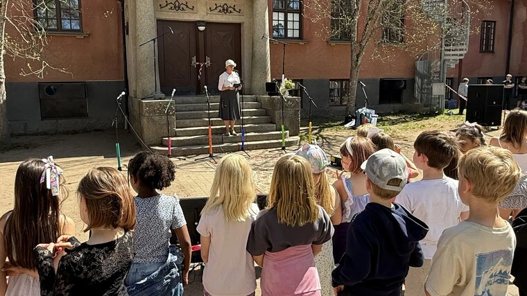 En grupp barn står samlade utomhus framför en byggnad med stora trädörrar, medan en person på trappan håller ett tal eller läser ur ett papper. Färgglada markeringar är placerade på marken framför trappan, och solen skiner över scenen.