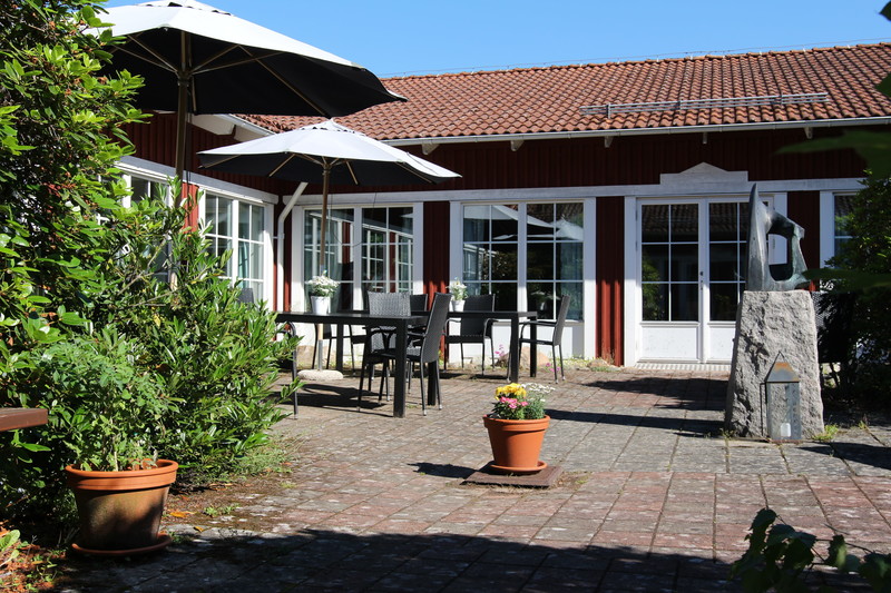 Uteplats med bord och stolar under stora parasoller, omgiven av grönska och med en röd byggnad med vita fönster i bakgrunden. Soligt väder med klarblå himmel.