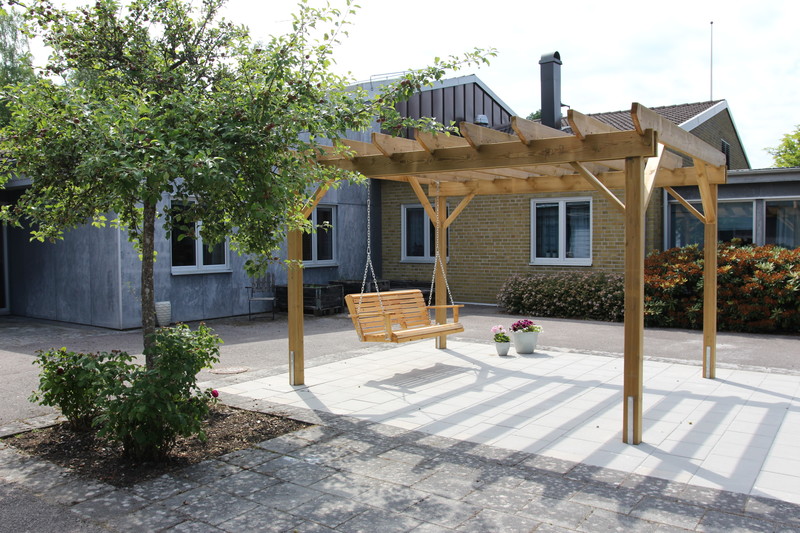 Träpérgola med gungbänk på stenlagd yta, omgiven av grönska och med byggnader i bakgrunden.