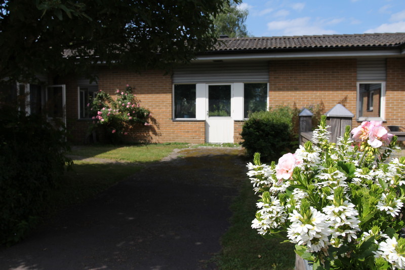 Enplans tegelbyggnad med mörkt tak och vit dubbeldörr vid entrén. Gångväg leder fram till dörren, omgiven av blomkrukor med vita och rosa blommor samt buskar. Delvis molnig himmel i bakgrunden.