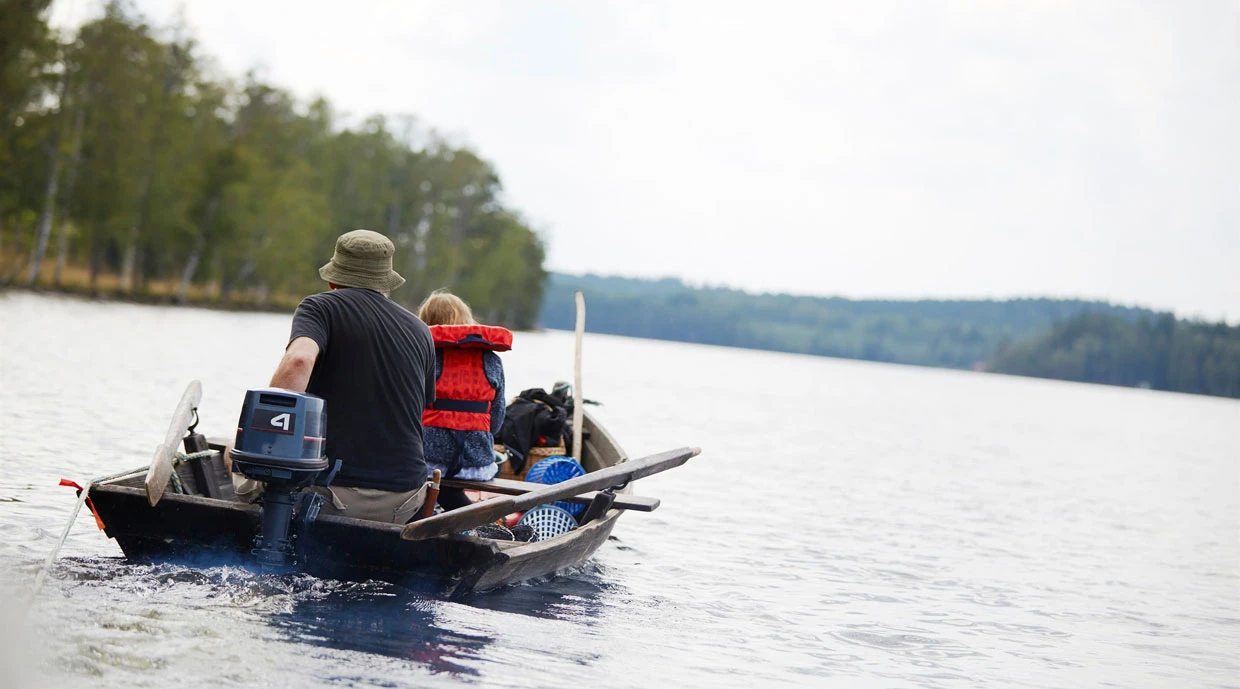 Två personer i en liten båt på en sjö, varav en bär hatt och mörka kläder och den andra en röd flytväst. Båten har en utombordsmotor och fiskeredskap ombord, med skog längs strandkanten i bakgrunden.