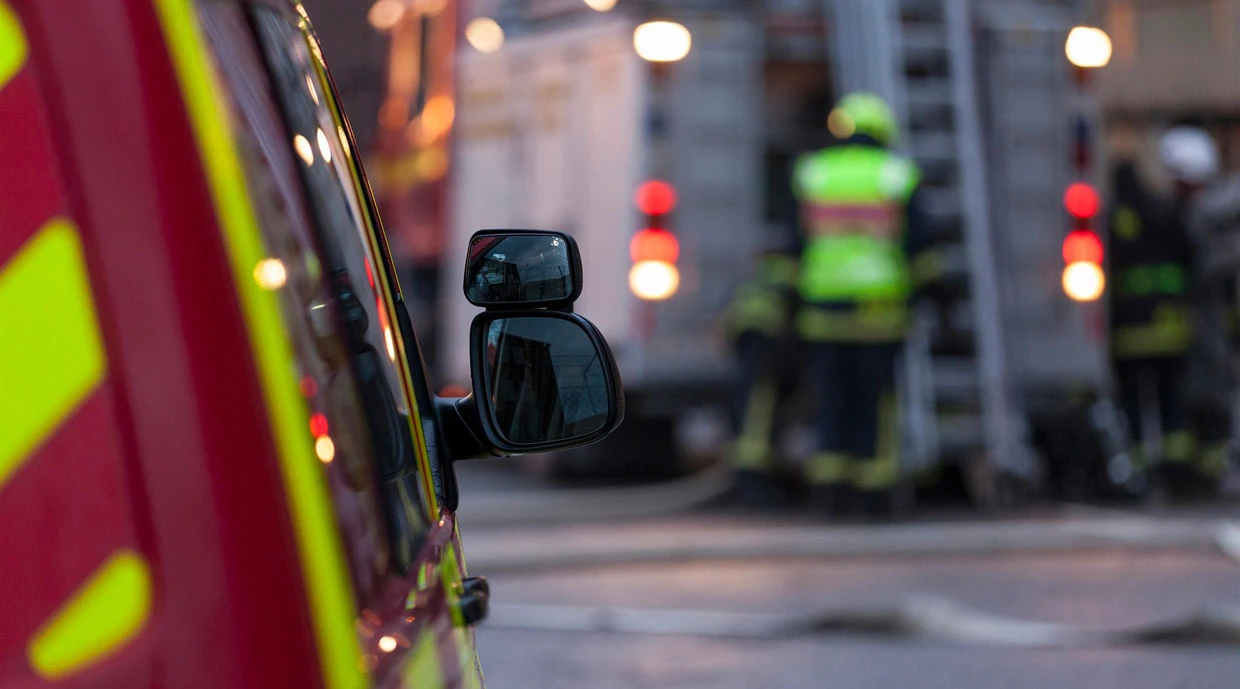 Räddningsinsats med brandmän i reflexkläder vid en röd och gul utryckningsfordon, med slangar på marken och aktivt arbete i bakgrunden.
