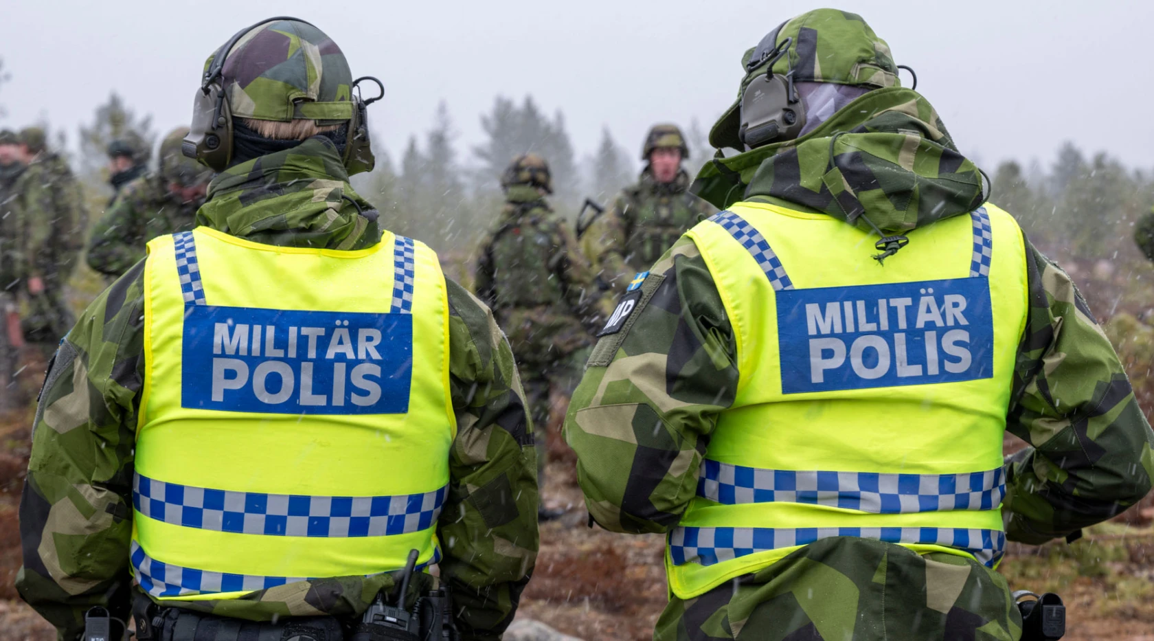 Två militärpoliser i kamouflageuniformer och gula reflexvästar med texten "MILITÄR POLIS" står med ryggen mot kameran. De befinner sig utomhus i en skogsmiljö tillsammans med andra personer i bakgrunden som också bär kamouflagekläder.