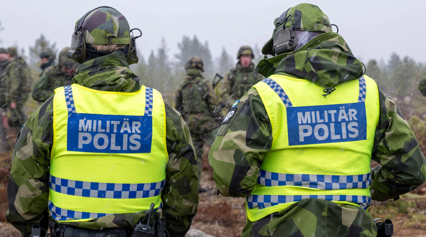 Två militärpoliser i kamouflageuniformer och gula reflexvästar med texten "MILITÄR POLIS" står med ryggen mot kameran. De befinner sig utomhus i en skogsmiljö tillsammans med andra personer i bakgrunden som också bär kamouflagekläder.