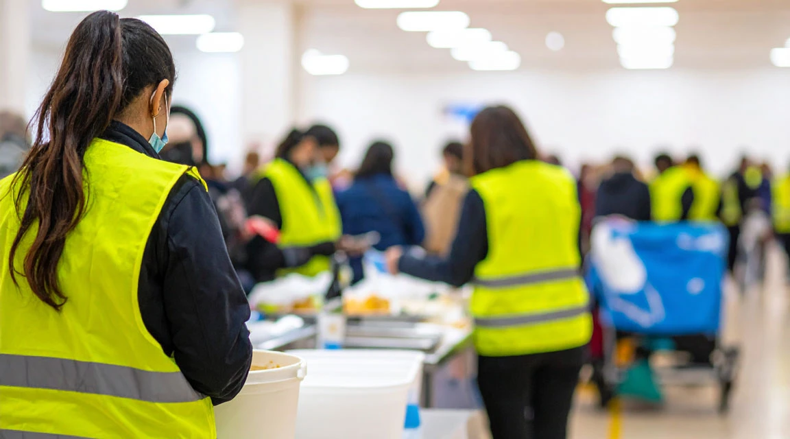 Grupp människor i gula reflexvästar arbetar tillsammans i en stor inomhuslokal, hanterar mat och andra föremål på bord – en samordnad insats med flera deltagare.