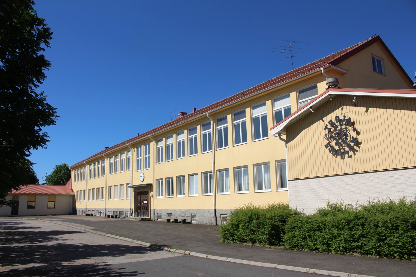 Gul skolbyggnad omringad av asfalt, buskar och träd.