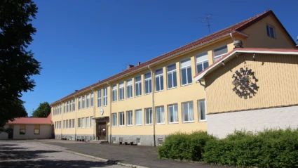 Gul skolbyggnad omringad av asfalt, buskar och träd.