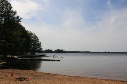 Badplats vid en lugn sjö med sandstrand i förgrunden, där leksaker som hinkar och spadar ligger utspridda. En träbrygga sträcker sig ut i vattnet med några personer stående på den. Träd ger skugga till vänster och himlen är delvis molnig.