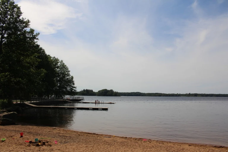 Badplats vid en lugn sjö med sandstrand i förgrunden, där leksaker som hinkar och spadar ligger utspridda. En träbrygga sträcker sig ut i vattnet med några personer stående på den. Träd ger skugga till vänster och himlen är delvis molnig.