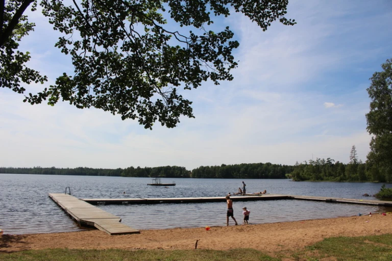 Strand vid en insjö med en brygga som sträcker sig ut i vattnet. Några personer syns på stranden och bryggan, och ett par badar. Skog omger sjön och himlen är blå med några moln.