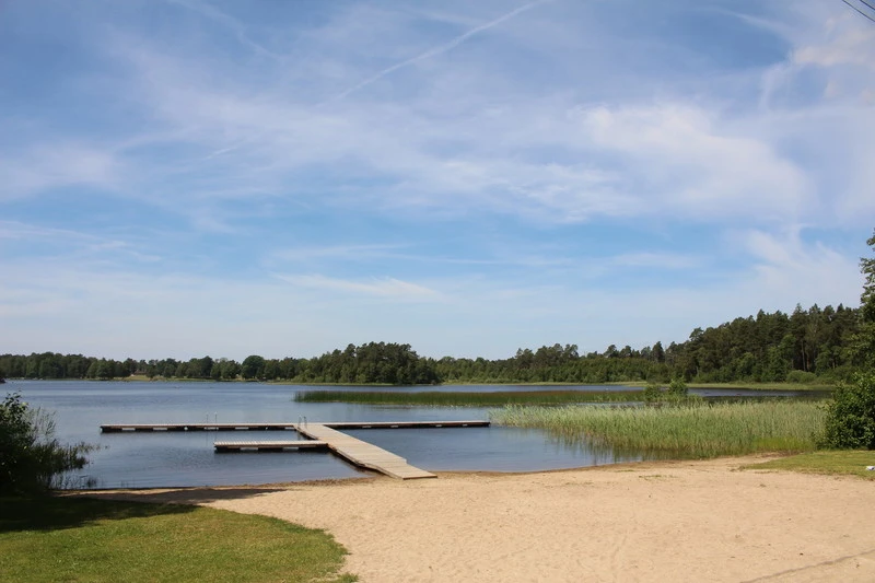 Brygga vid en sjö med sandstrand i förgrunden och tät skog i bakgrunden.