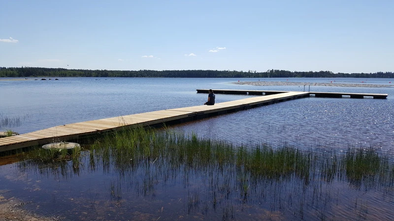 Träbrygga i T-form som sträcker sig ut i en stilla sjö, med en person sittande vid kanten och blickande ut över vattnet. Klarblå himmel, skog i bakgrunden och vass vid strandkanten.
