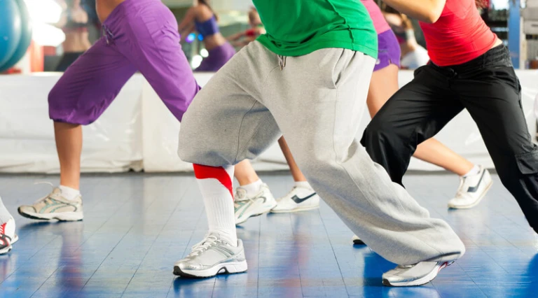 En grupp personer deltar i ett träningspass på gymmet, klädda i färgglada träningskläder och sneakers, medan de utför dansrörelser på ett blått golv.