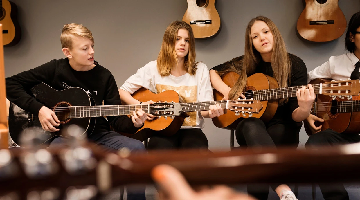 Tre personer sitter i en musikklass och spelar akustiska gitarrer. Flera gitarrer hänger på väggen bakom dem, och en gitarr i förgrunden är delvis synlig.