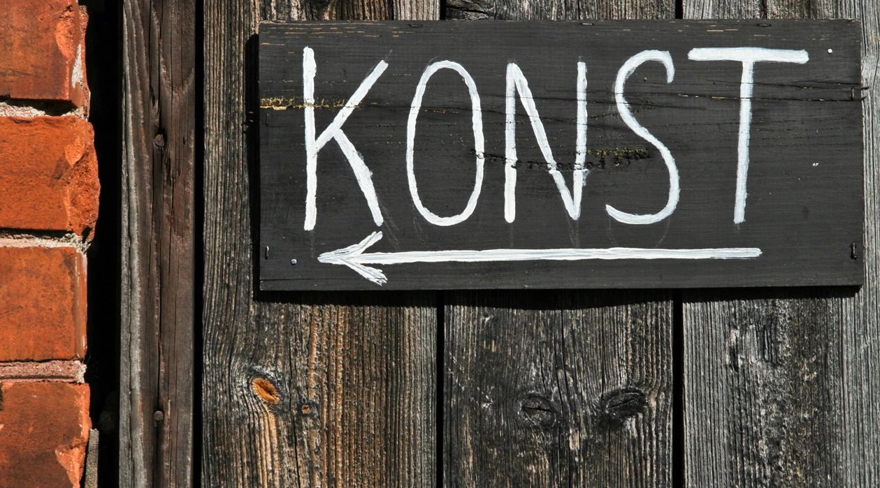 Svart träskylt med vit text som säger ”KONST” och en pil som pekar åt vänster, monterad på en vägg av väderbitet trä bredvid en tegelvägg.