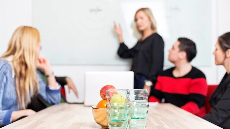 En grupp personer sitter vid ett konferensbord med glas och frukt i förgrunden, medan en person står vid en whiteboard och presenterar något för de andra.