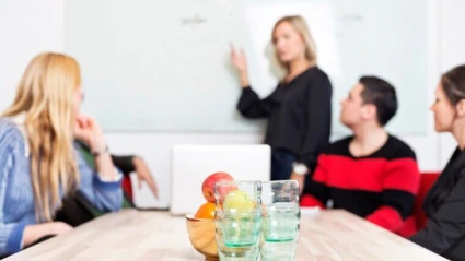 En grupp personer sitter vid ett konferensbord med glas och frukt i förgrunden, medan en person står vid en whiteboard och presenterar något för de andra.
