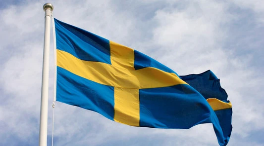Svenska flaggan med blå bakgrund och gult kors vajar mot en ljus himmel med moln.