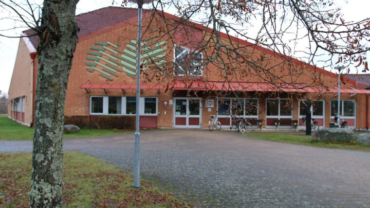 Wasaskolans entré till en tegelbyggnad med rött tak, stora fönster och en gångväg som leder fram till dörrarna. Några cyklar står parkerade utanför, och träd med höstlöv ramar in bilden.