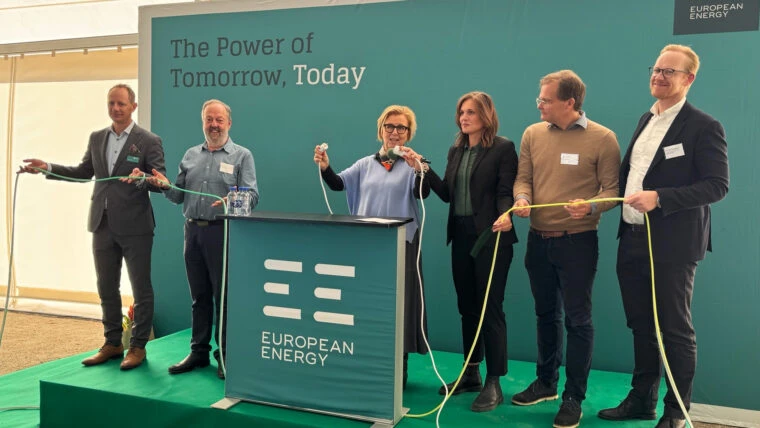 En grupp personer står på en scen framför en grön bakgrund med texten ”The Power of Tomorrow, Today” och logotypen för European Energy. De håller i långa kablar som en del av en ceremoni, med ett podium i mitten.