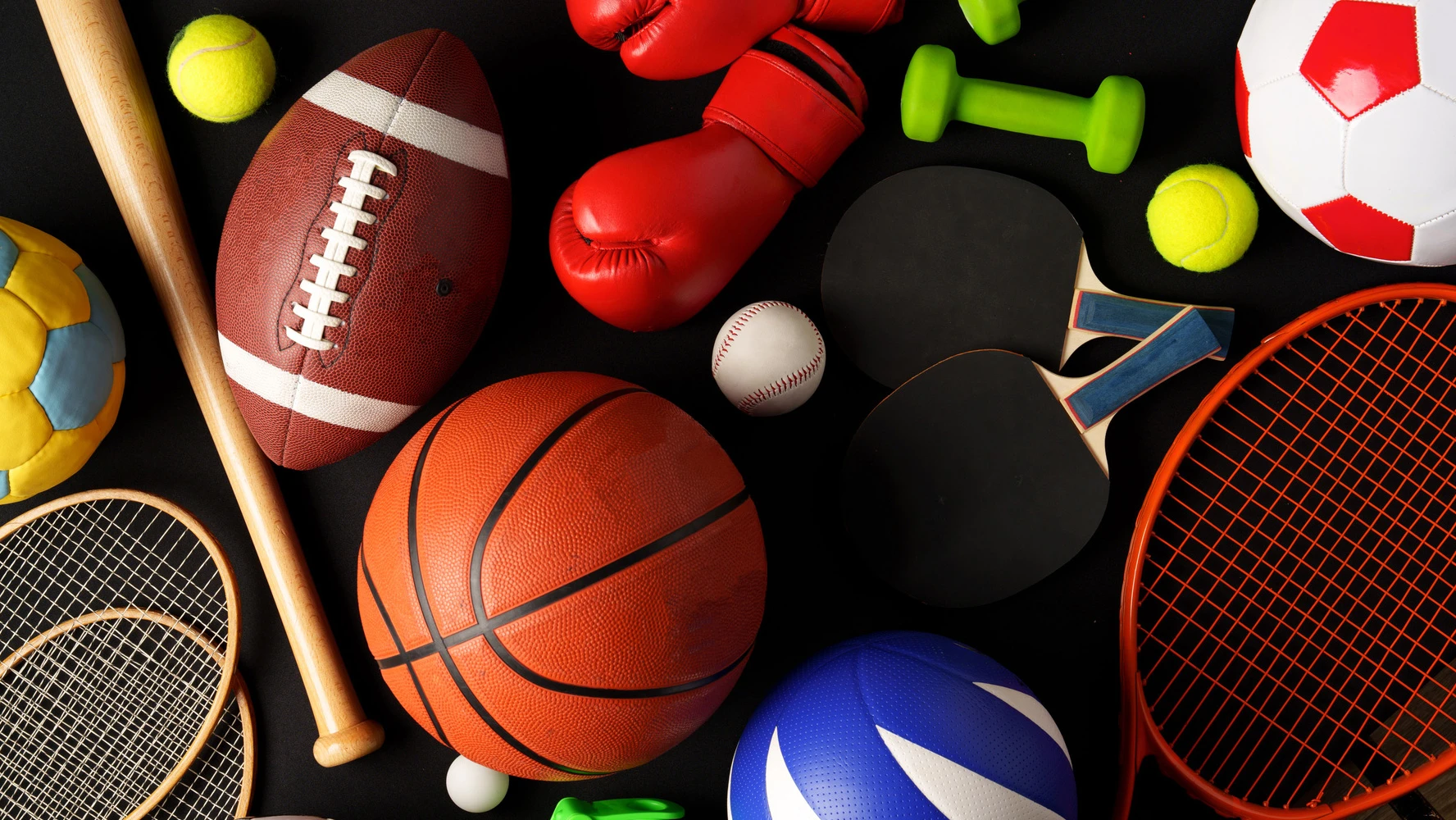 Olika sportutrustningar arrangerade mot en svart bakgrund, inklusive en fotboll, basketboll, tennisracket med boll, basebollträ med boll, amerikansk fotboll, boxningshandskar och en hopprep. Bilden är tagen i studio med jämn belysning som framhäver detaljerna på utrustningen.