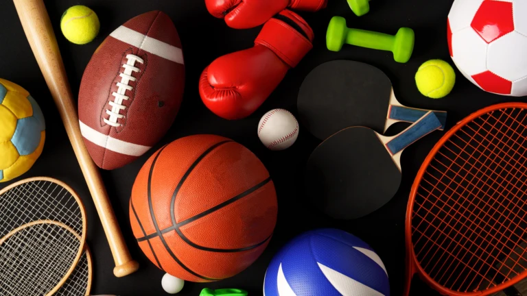 Olika sportutrustningar arrangerade mot en svart bakgrund, inklusive en fotboll, basketboll, tennisracket med boll, basebollträ med boll, amerikansk fotboll, boxningshandskar och en hopprep. Bilden är tagen i studio med jämn belysning som framhäver detaljerna på utrustningen.